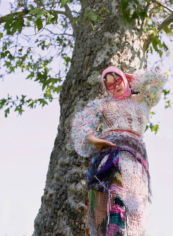 Woman in elaborate crocheted dress and pink headscarf poses next to a large tree with sparse leaves against a bright sky.