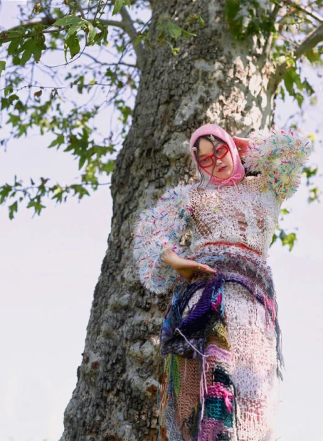 Woman in elaborate crocheted dress and pink headscarf poses next to a large tree with sparse leaves against a bright sky.