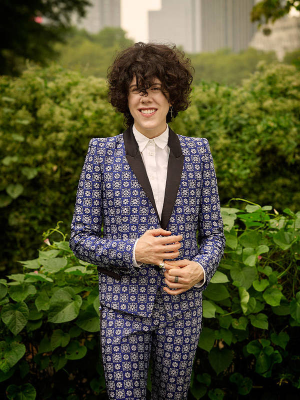 LP, a singer-songwriter, stands in patterned blue suit among greenery in an eye-level outdoor portrait with buildings visible in background.