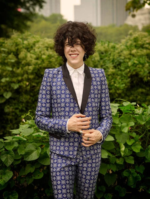 LP, a singer-songwriter, stands in patterned blue suit among greenery in an eye-level outdoor portrait with buildings visible in background.