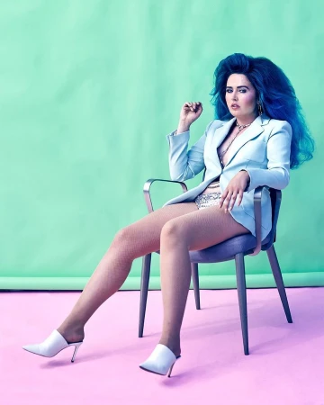 Portrait of a woman with blue hair sitting on a chair, wearing a light blue blazer, shiny shorts, white heels against a gradient backdrop.