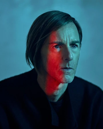 Portrait of a fair-skinned adult male looking slightly off-center, with a red light bisecting his face, wearing a black sweater.
