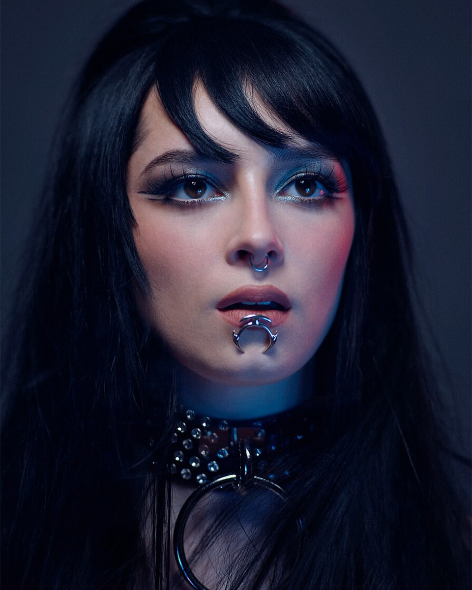 Close-up portrait of a young adult woman with dark hair, silver lip ring and nose ring, wearing a studded choker necklace.