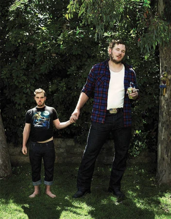 A playful image of two men, one notably taller, holding hands in a lush backyard setting, under the shade of leafy trees.