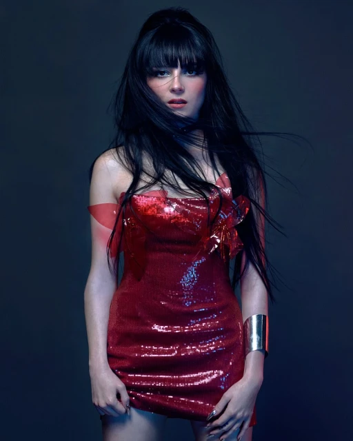 A fair-skinned woman with black hair poses indoors wearing a red sequin dress with a silver bracelet, against a cool, shadowy background.