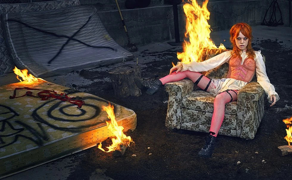 Edgy fashion portrait of a woman in a pink corset and thigh-highs sitting in an armchair surrounded by burning mattresses in an outdoor setting.