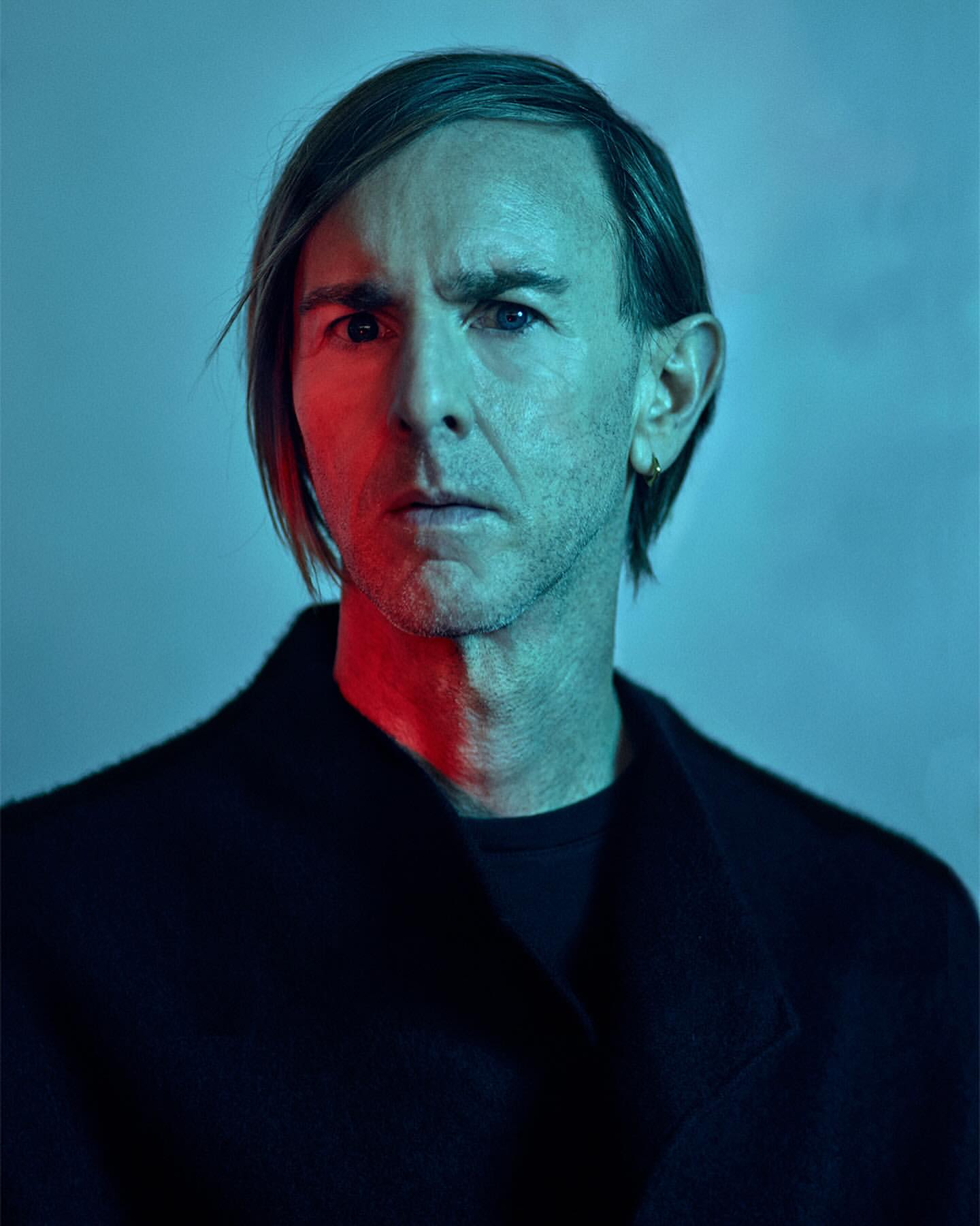 Portrait of a light-skinned adult man with short hair wearing a black overcoat. He is posing against a blue backdrop with red lighting.