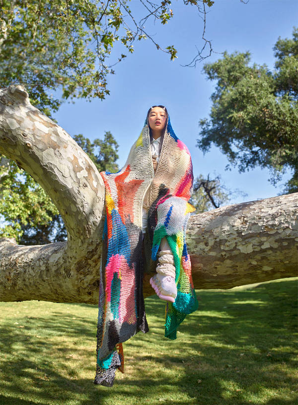 An Asian woman is sitting on a tree branch wearing a colorful, draped shawl in an outdoor park setting on a bright sunny day.