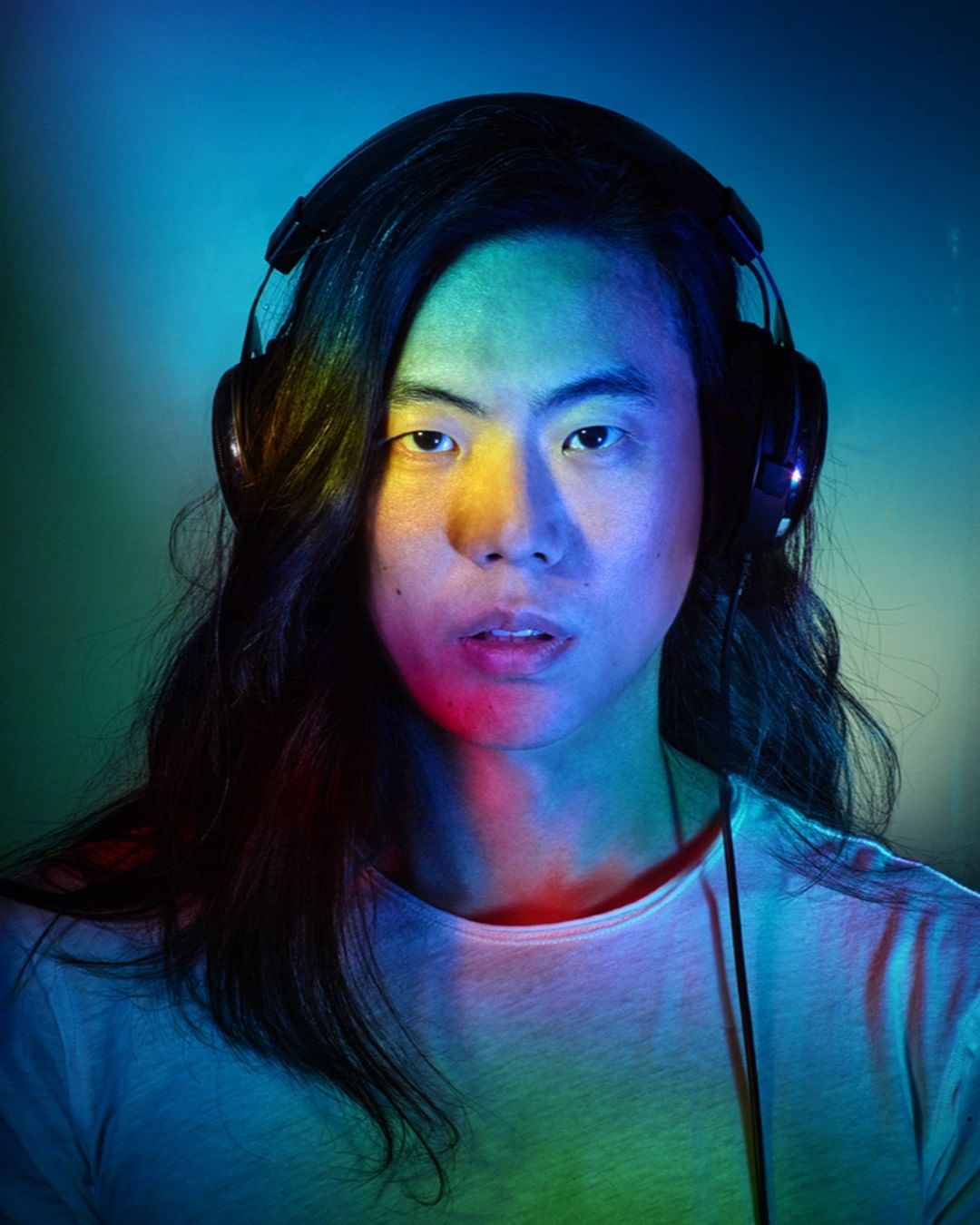 Close-up portrait of an Asian adult with long black hair, wearing headphones and a white shirt, illuminated by multicolored lights.