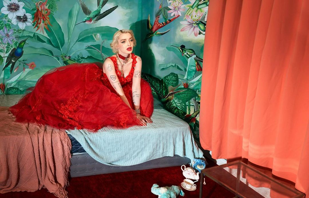Woman in red gown sits on bed in room with floral wallpaper and coral drapes. Interior with objects and props. Bright lighting and color.