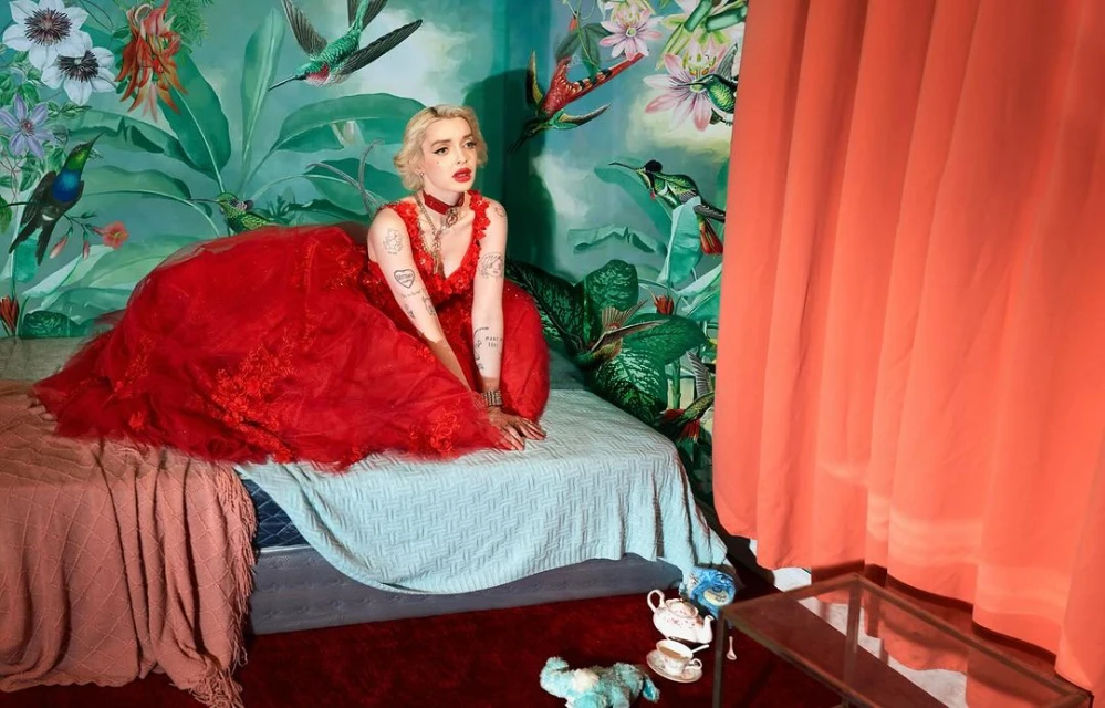 Woman in red gown sits on bed in room with floral wallpaper and coral drapes. Interior with objects and props. Bright lighting and color.