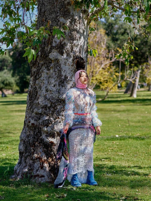 Young woman in a hooded dress posing by a tree in a park setting on a sunny day, showcasing a unique fashion style and natural outdoor scene.