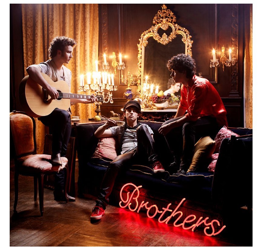 Three young men inside ornate room: one sitting on chair playing guitar, one on sofa with a hat, one sitting on the back of the sofa.