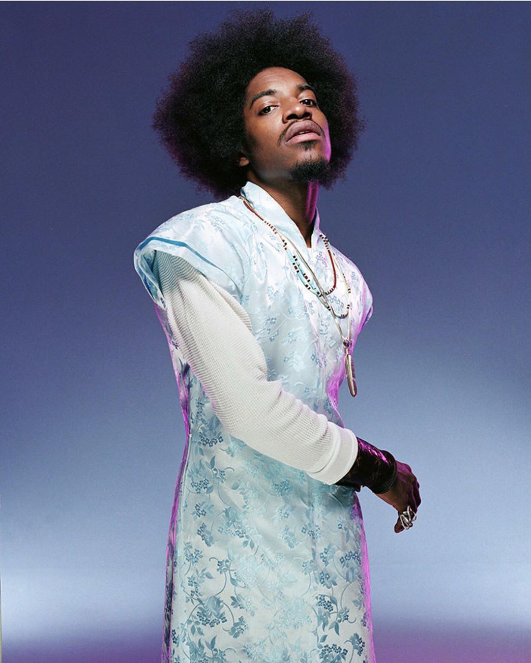 Studio shot of a Black male model with an afro, wearing a light blue patterned tunic, beaded necklaces, and a dark bracelet.