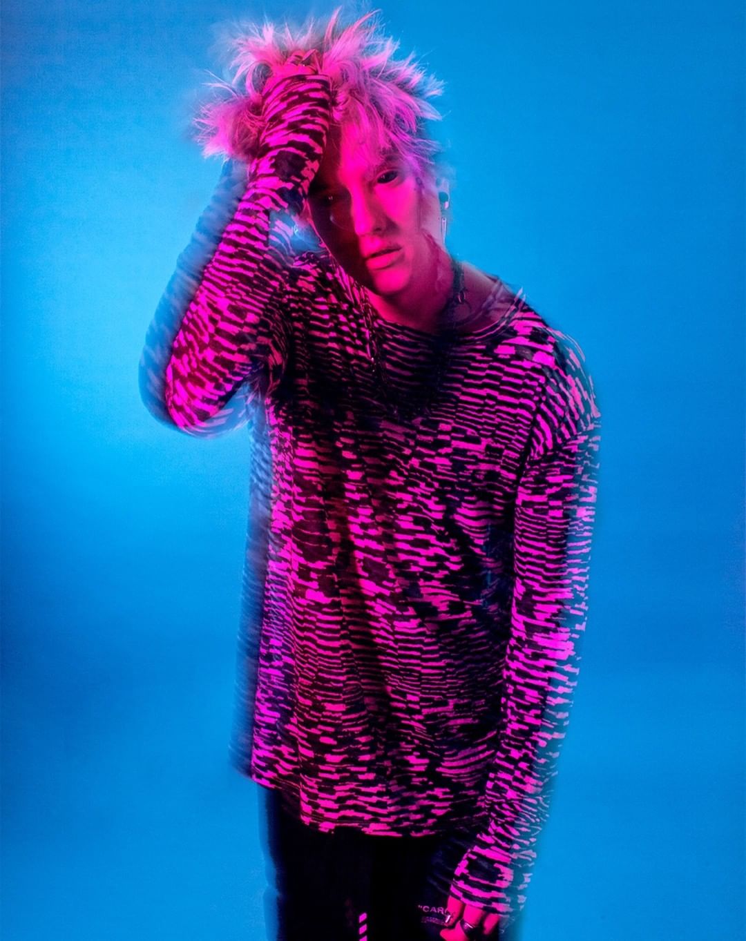 Portrait of a person with pink-tipped blond hair wearing a patterned shirt against a blue background, gazing intently at the camera.