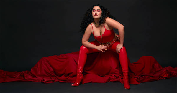 Brunette adult woman poses in a red bodysuit, leggings, and high heels with a red fabric and a black backdrop in a studio setting.