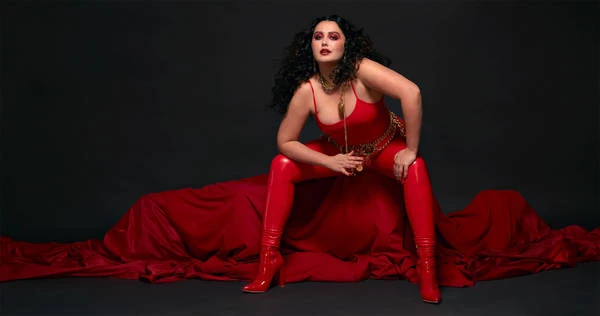 Brunette adult woman poses in a red bodysuit, leggings, and high heels with a red fabric and a black backdrop in a studio setting.