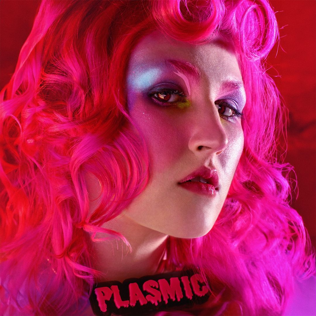 Close-up portrait of a woman with hot-pink curly hair and bold colorful makeup against a red background wearing a name badge.