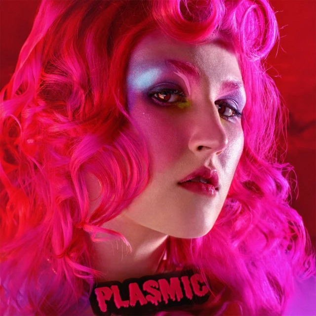 Close-up portrait of a woman with hot-pink curly hair and bold colorful makeup against a red background wearing a name badge.