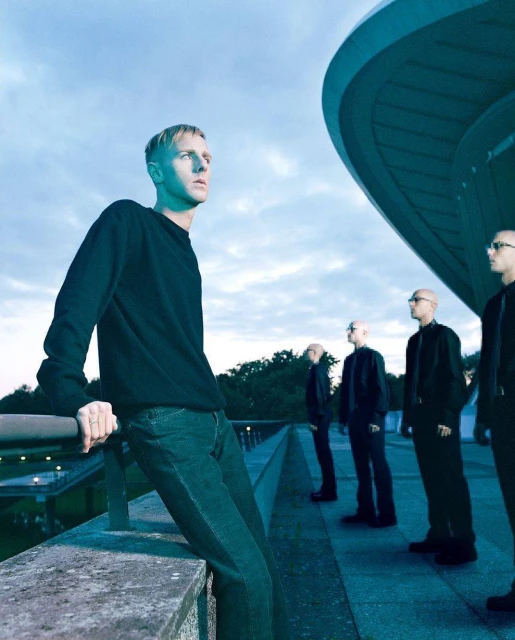 Group of men in dark clothing stand in an urban setting with modern architecture and moody lighting, looking in the same direction.