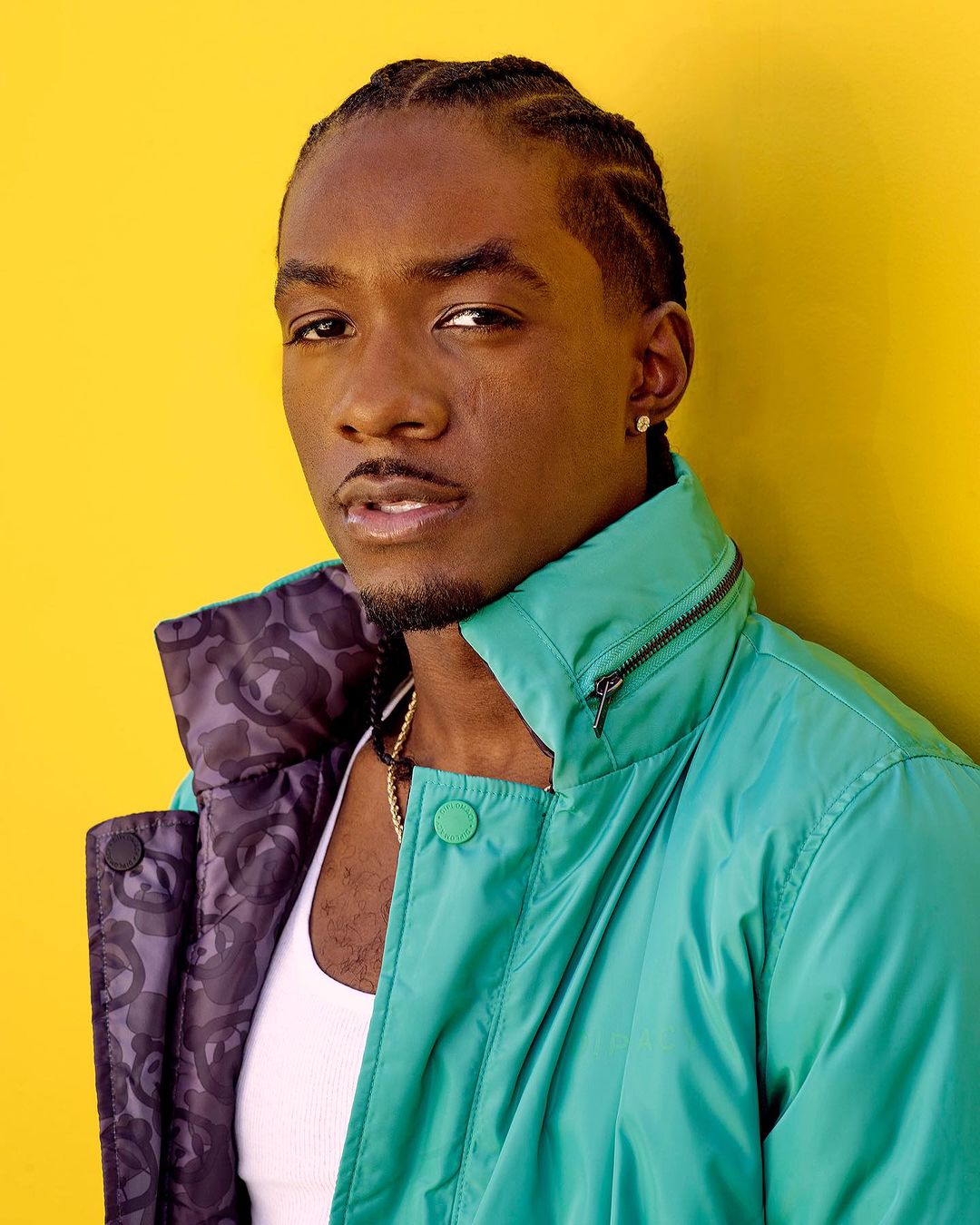 Portrait of an adult Black male with braided hair, wearing a teal jacket in front of a yellow backdrop, showcasing a clean and stylish look.