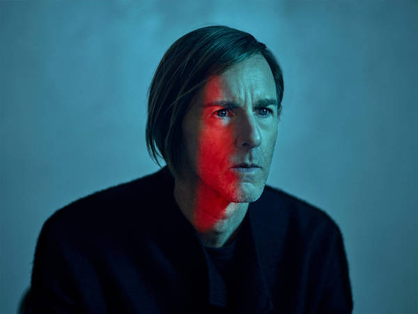 Portrait of a man in his 40s with side-parted hair wearing a dark jacket under blue and red studio lighting