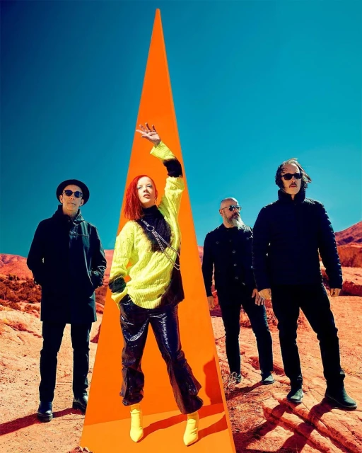 Four people stand in a desert landscape under a bright blue sky next to an orange triangular object. Fashionable clothing and striking colors.