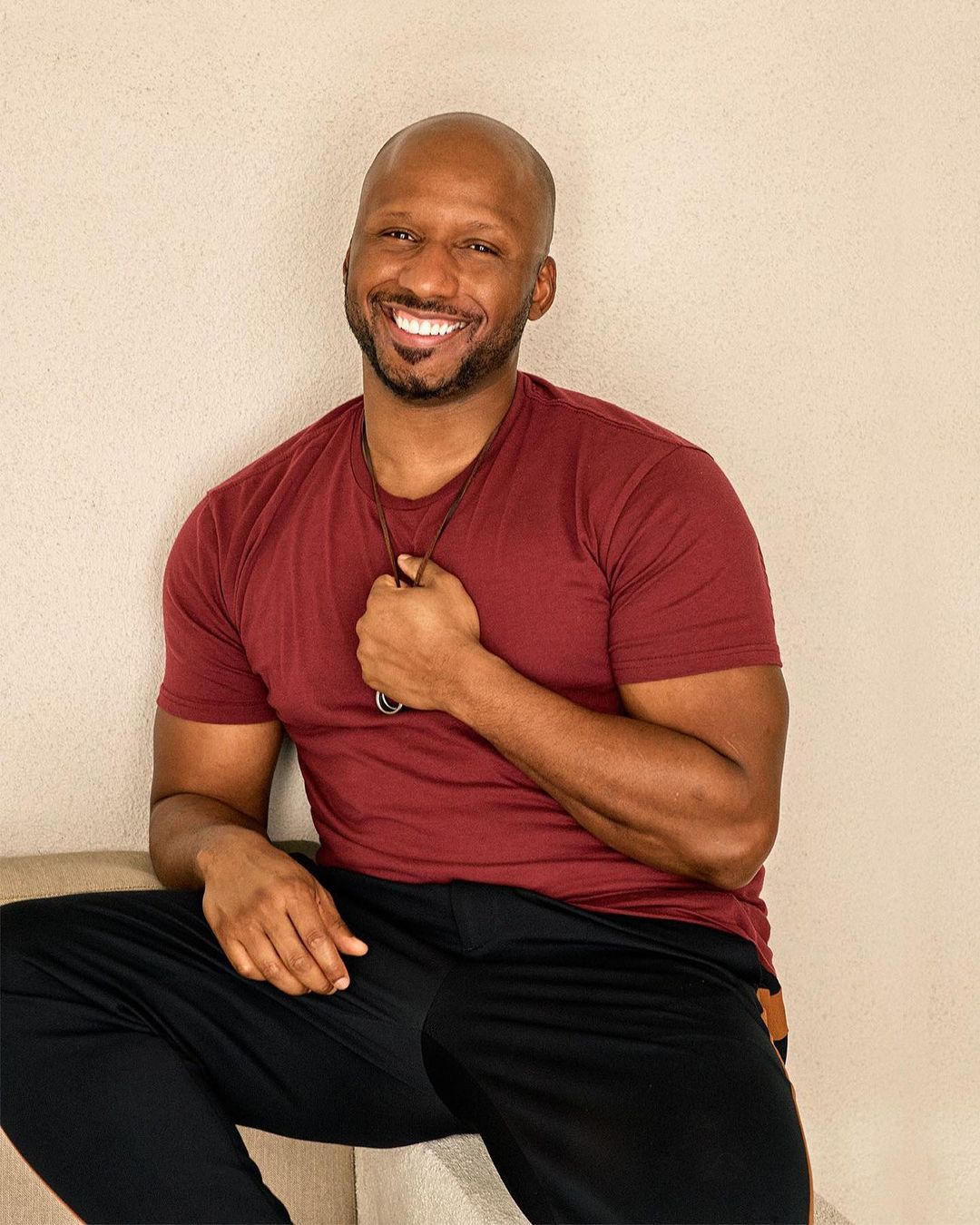 Smiling adult man with shaved head, wearing a maroon t-shirt and black pants, poses indoors with a light beige background.