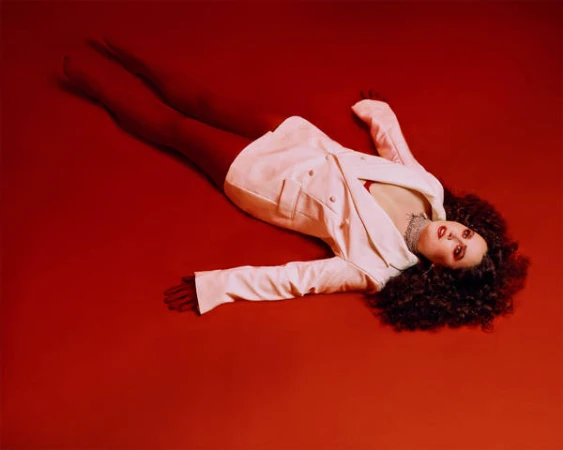 An adult woman with curly brown hair lies on a red surface, wearing a white jacket, red top, and dark red tights in a studio shot.