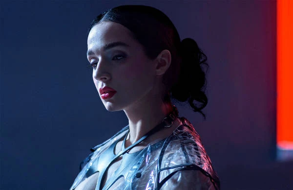 Portrait of a woman with dark hair in a low ponytail wearing a translucent plastic garment with black harness details under neon lighting