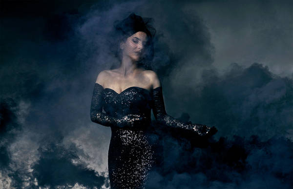 Woman in black dress with black lace gloves poses in a dramatic studio shot surrounded by dark smoke against a blue-gray background.