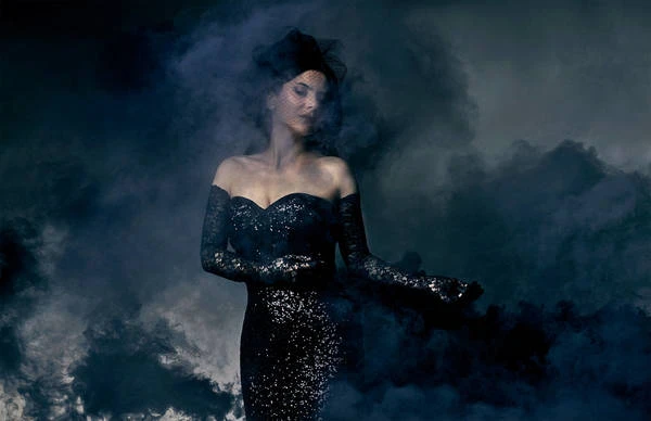 Woman in black dress with black lace gloves poses in a dramatic studio shot surrounded by dark smoke against a blue-gray background.