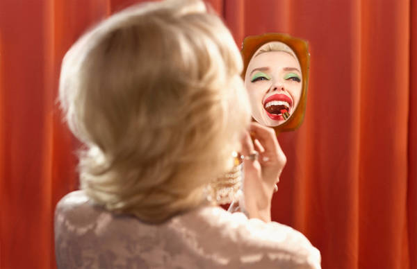 Blonde woman holding a hand mirror showing her reflection applying red lipstick against red curtains