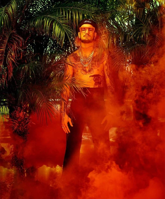Tattooed male model poses shirtless amid palm trees and orange smoke, wearing cap, glasses, pants, jewelry, and standing outdoors.