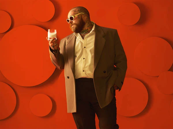 Adult male with beard and tattoos wearing sunglasses and suit jacket, holding a glass of milk in a studio with an orange background.