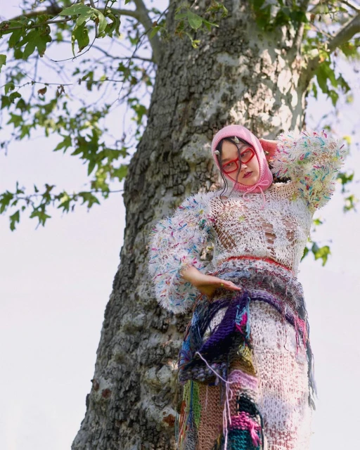 An adult female stands outside near a tree, wearing a crocheted outfit and pink headwear, in a whimsical fashion style.