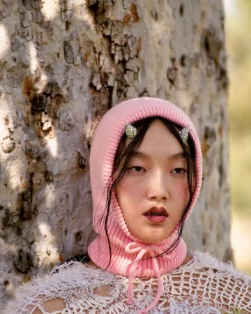 Close-up of an Asian woman wearing a pink knitted balaclava and decorative hair clips, with a light beige tree trunk in the background.