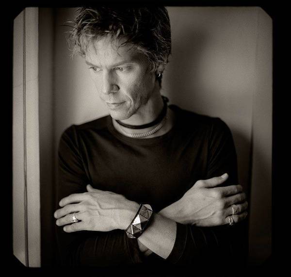 Portrait of an adult male with fair skin and short spiky hair wearing a black shirt and jewelry, looking down with arms crossed.