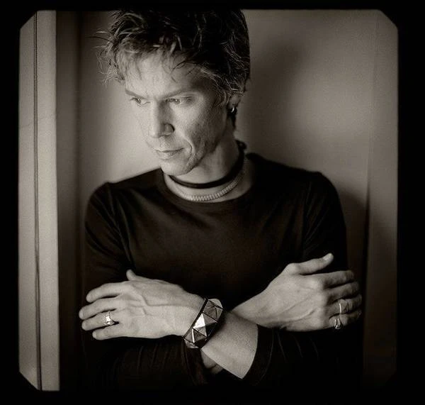 Portrait of an adult male with fair skin and short spiky hair wearing a black shirt and jewelry, looking down with arms crossed.