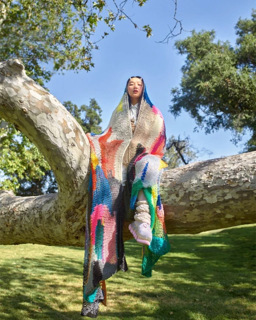 An Asian woman sits on a tree branch wearing a colorful knitted wrap, posing in a park-like setting with green grass and trees under a blue sky.