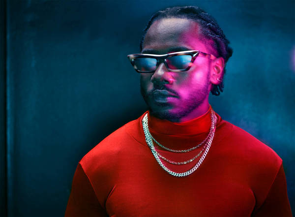 Portrait of a black man wearing sunglasses, a red turtleneck, and silver chain necklaces under blue and pink lighting.