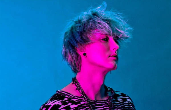 Profile portrait of a young adult with dyed hair and chain necklace, illuminated by bright blue and pink studio lighting.