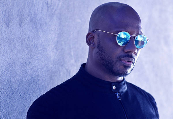 Headshot of a bald adult male with a beard wearing mirrored sunglasses, a black jacket, and standing against a textured wall.