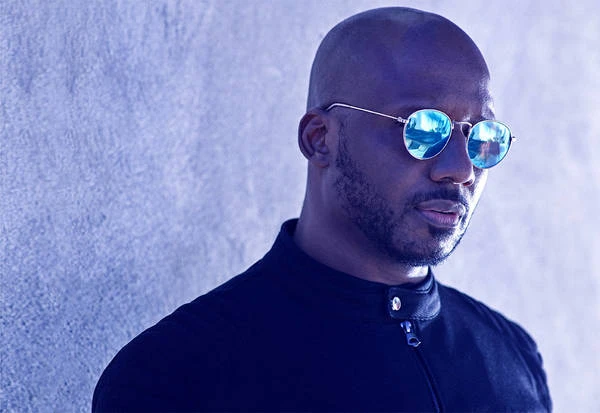 Headshot of a bald adult male with a beard wearing mirrored sunglasses, a black jacket, and standing against a textured wall.