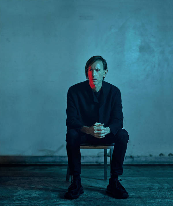 Man seated on a chair against a textured blue and red lit concrete wall wearing dark clothing by Joseph Cultice