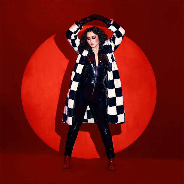Woman posing in shiny jumpsuit and checkered coat against a red backdrop with a circular light in a fashion or music setting.