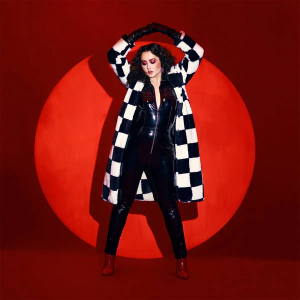 Woman posing in shiny jumpsuit and checkered coat against a red backdrop with a circular light in a fashion or music setting.