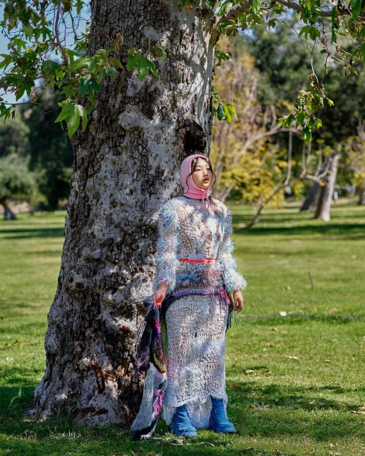 Young woman in knitwear fashion standing by a tree in a park, wearing a pink headscarf and blue boots. Sunlight and natural setting.