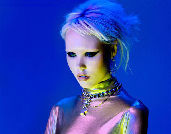 Close-up studio portrait of a young adult woman with platinum blonde hair, wearing bold eye makeup and multiple necklaces against a blue backdrop.
