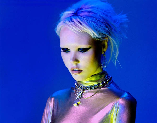 Close-up studio portrait of a young adult woman with platinum blonde hair, wearing bold eye makeup and multiple necklaces against a blue backdrop.